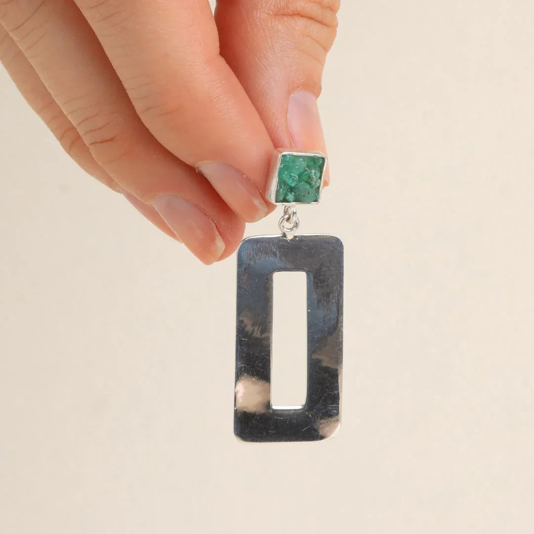 Hand holding a silver rectangular earring with a raw green emerald stud.