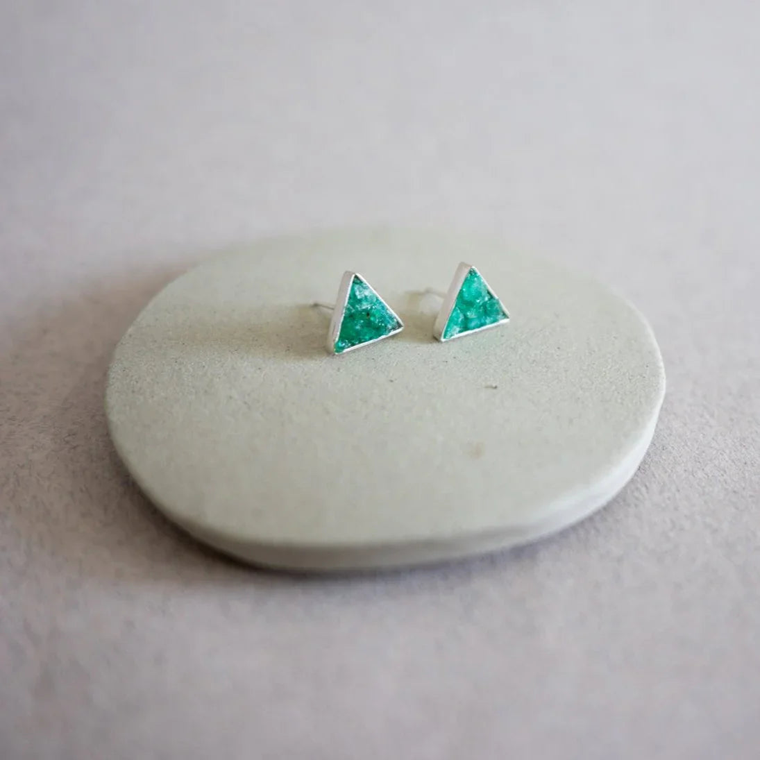 Silver triangular stud earrings with vibrant green Colombian emeralds, displayed on a light gray disc.