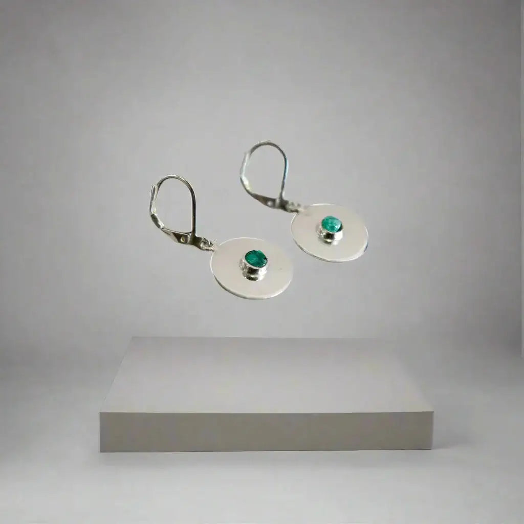 Handmade recycled silver drop earrings with small round Colombian emeralds, floating above a gray pedestal.