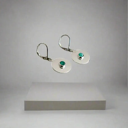 Handmade recycled silver drop earrings with small round Colombian emeralds, floating above a gray pedestal.