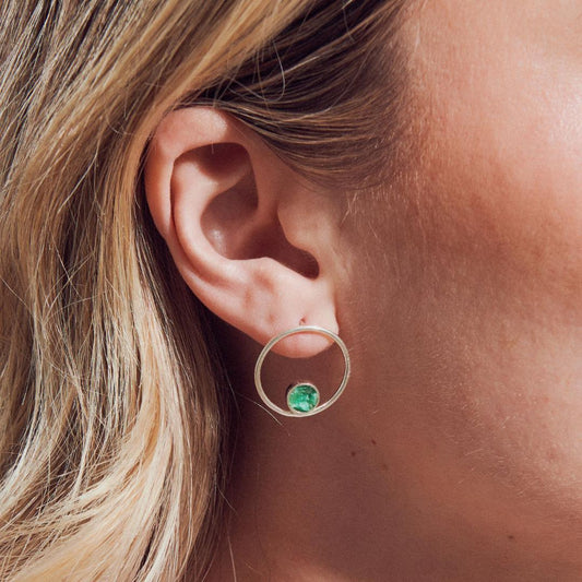 Silver hoop earring with a vibrant green emerald, worn on a woman's ear.
