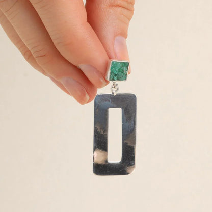 Hand holding a silver rectangular earring with a raw green emerald stud.