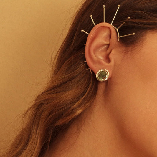 Silver ear cuff with sunburst spikes and a round emerald stud earring on a woman's ear.