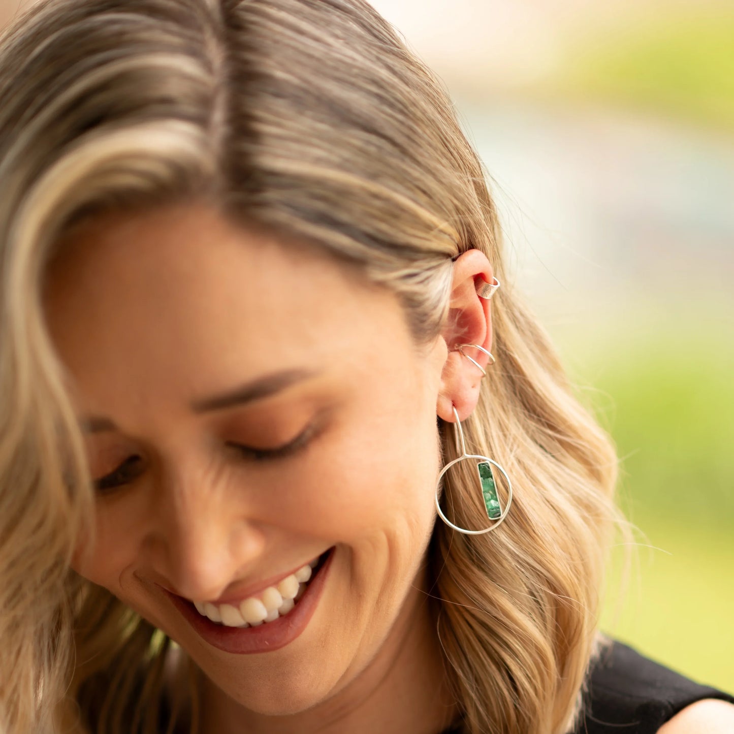 Woman wearing a silver hoop earring with a rectangular emerald bar, and multiple silver ear cuffs.